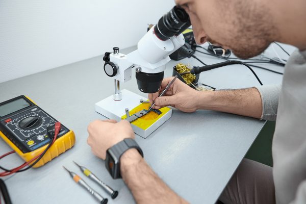 Cropped view of skilled young bearded male technician carrying out microscopic examination of cell phone small parts. Repairman examining mobile phone motherboard under microscope in laboratory